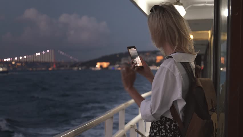 Young woman taking pictures of sea and coastal city while traveling on water by boat or ferry in evening or at night. Lights of city are burning. Using camera on your smartphone during boat cruise