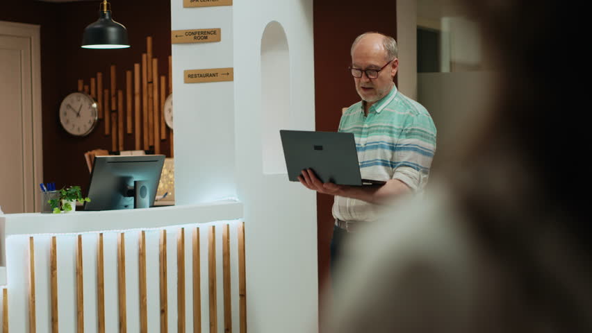 Senior people in hotel reception lobby, woman arriving at front desk with luggage while man is waiting in lounge area using laptop. Receptionist assisting travellers at luxury resort. Handheld shot.