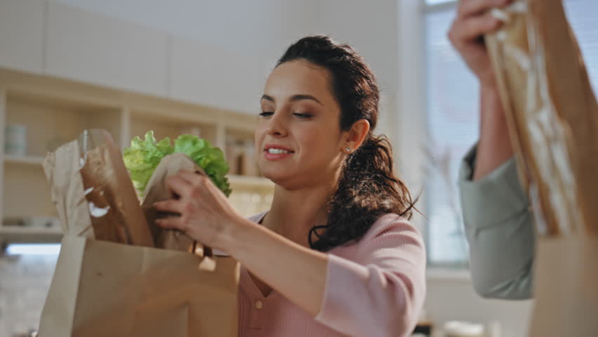 Closeup family taking purchased products on kitchen countertop from paper bags. Happy cheerful couple unpacking ecological supermarket packages with healthy food. Spouses sorting shoppings together.