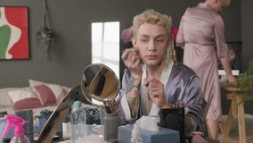 Two beautiful young Caucasian queer men in silk robes applying makeup while getting ready together in cluttered apartment at daytime