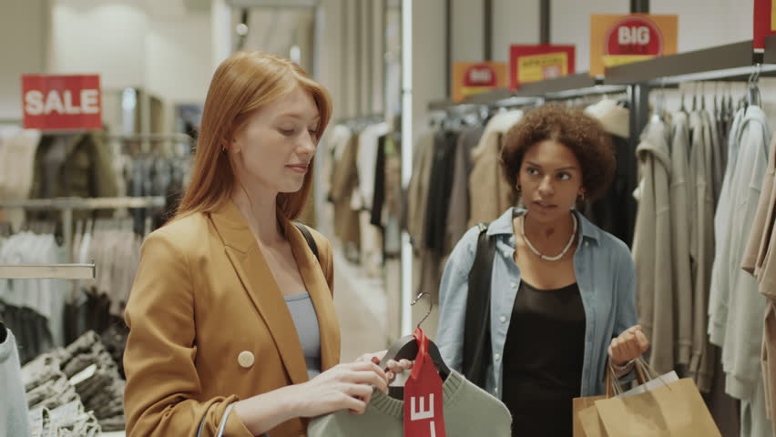 Medium shot of two young multiracial women pulling clothing item and arguing while doing shopping in clothes store during seasonal discounts