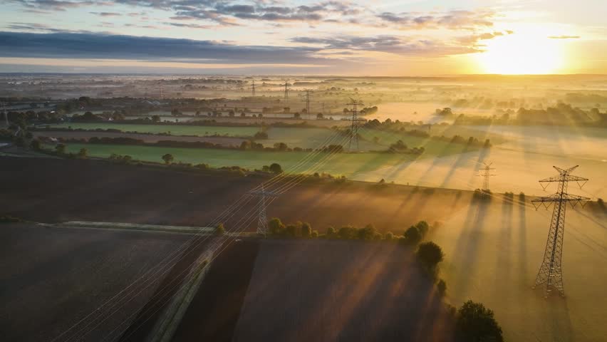 Morning sunrise over industry utility power lines and pylon structure in foggy agricultural landscape as the camera swings from left to right. Multiple power lines and pylons stretch the farmland. Royalty-Free Stock Footage #1110097477