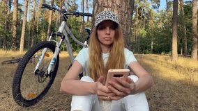 A girl shares news via smartphone on the Internet with friends, discussing cycling. Portrait of a happy female cyclist sitting on a meadow in the forest near a tree, resting. High quality 4k video