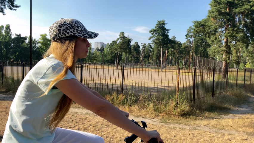 A girl on a white bicycle rides along the road along the stadium and trees. A breezy mountain bike ride. Fresh air in the forest. A girl relaxes on her bike, pedaling along the pine trees. High 4k