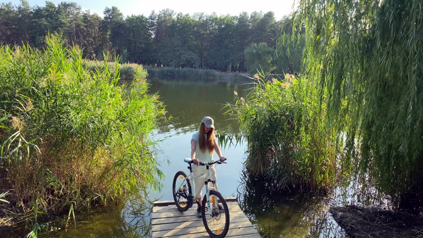 Young woman on a wooden bridge with a bicycle near the lake. The girl is standing on a wooden block, holding a white bicycle in her hands, a sunny summer day near the lake. High quality 4k video