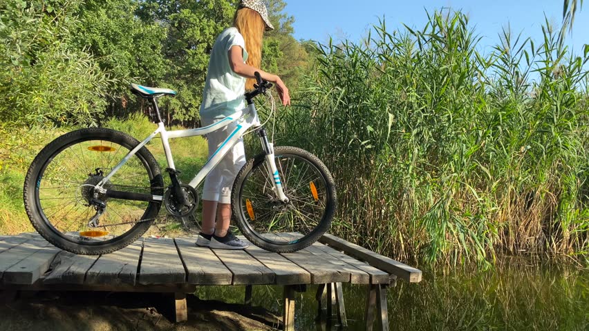 By the lake, a girl on a bicycle looks into the water, standing on a bridge. A woman by the water watches the swimming fish while holding a mountain bike. Around reeds and green bushes. 4k