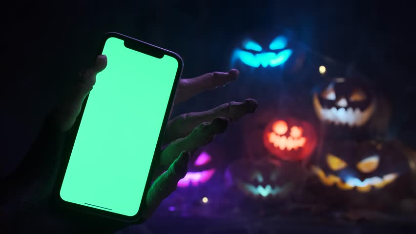 Skeleton hand holds a phone with chroma key screen against the background of colorful flashing Jack-o'-lanterns sends out an invitation to Halloween party during holiday.