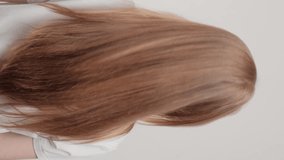 Vertical video. A red-haired girl stands with her back in the studio on a gray background and waves her hair. The concept of caring for beautiful and healthy hair.