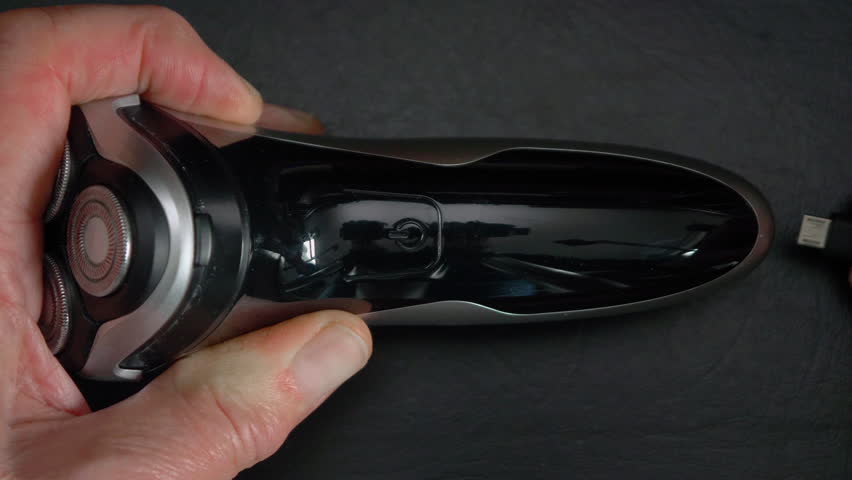 Closeup of a man’s hands holding a battery powered shaver while plugging in the charger cord, which then activates the charging display.