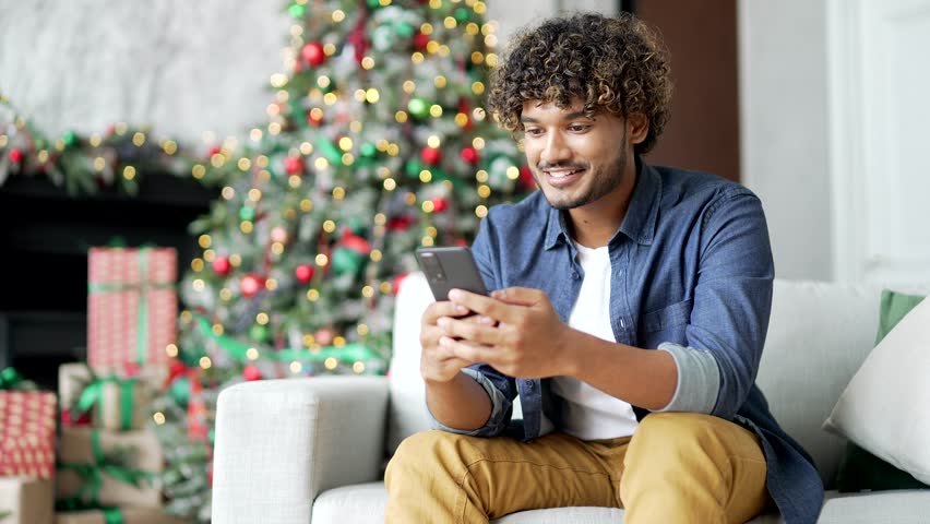 Close up. A young man uses a smartphone while sitting on sofa at home during winter New Year Christmas holidays. A handsome male is writing a greeting message, chatting with friends or shopping online Royalty-Free Stock Footage #1110113435