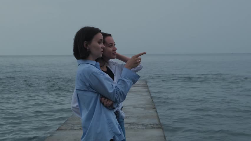 Two girls standing on breakwater and looking at sea horizon. Girlfriends enjoying seascape panorama with calm waves and having conversation