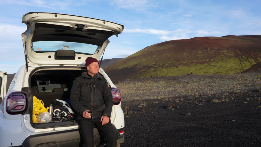 A man sits on the luggage compartment of his parked car, sipping coffee from a cup and enjoying the view near the Raudhaskal crater