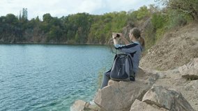 Middle-aged woman sits on elevated rocky shore and takes video on her smartphone. Hiking tourism concept.