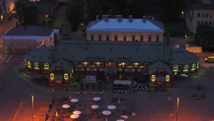 Aerial slide and pan footage of historic building of Hietalahti market hall in evening. Square lit by streetlights. Helsinki, Finland