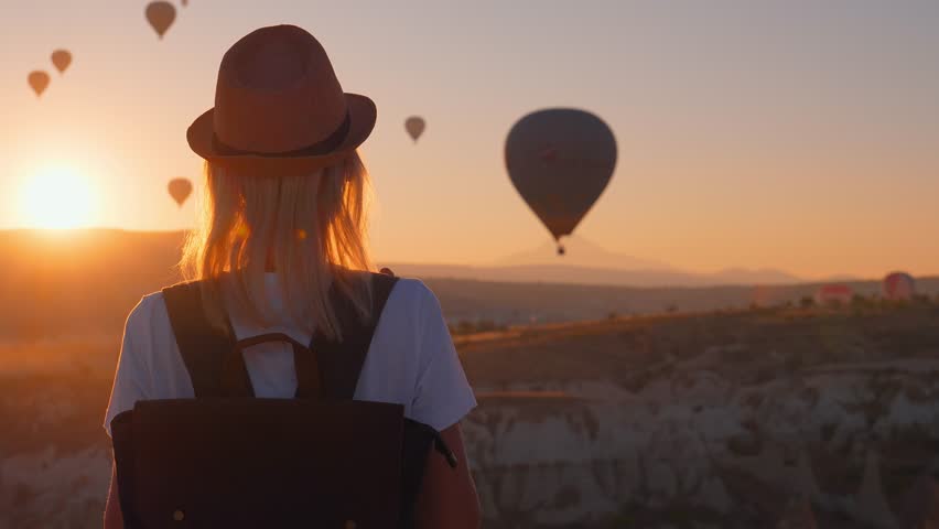 Young beautiful blonde woman traveler with backpack and hat walking and enjoying hot air balloon rides on Goreme hill, Cappadocia, Turkey at dawn in sunshine of girl. Hiking in fairy tale Royalty-Free Stock Footage #1110128217