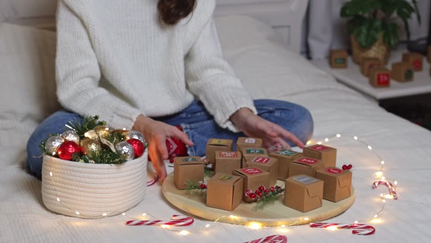 Hands of a caucasian teenage girl in a white knitted sweater and jeans are twisting a round tray with an advent calendar box and a burning garland, sitting on a bed, sitting on a bed in a room, close-