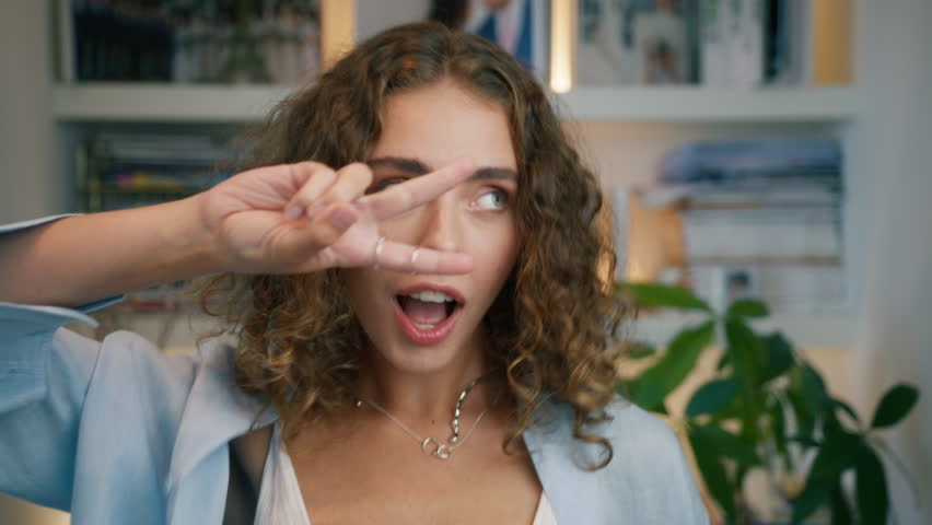 Young natural beauty woman with curly hair wearing blu linen shirt and white top dancing showing V sign 80s disco moves, fool around, have fun, enjoy weekend, celebrate, posing in apartment background