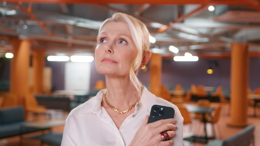 Concerned business woman with smartphone in hands looking around considering partnership offer or job opportunity, thinking through partnership terms, business agreement. Portrait mature elegant lady