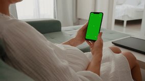 Pregnant female sitting on a sofa and taps on green screen phone in her hands. Vertical green screen phone hold by pregnant.