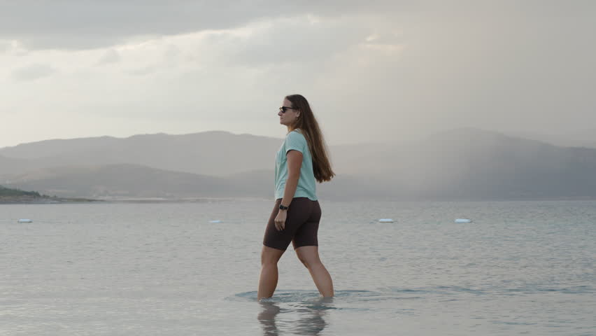 Approaching Storm Clouds in the distance near the mountains. They are moving towards the lake where a Young Woman is walking in the water and pointing at them.