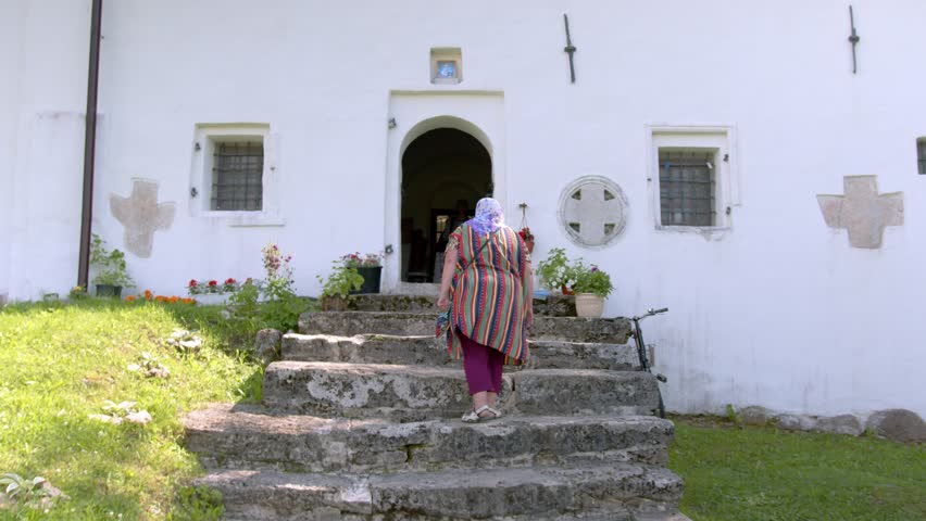 A Slavic adult village woman walks up the stairs to the doors of the village church and is baptized on the threshold. Religion and prayer. Life of the village community. Slavic modern villages