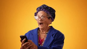 Toothless funny old grandmother wearing glasses and boa having video chat with family and friends isolated on yellow background. Concept of crazy senior youthful woman.
