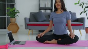 Young asian woman practicing yoga with trainer via video conference. Girl in lotus pose while sitting on mat in the morning at home. Relaxation and meditation Sporty athletic fitness workout sport.