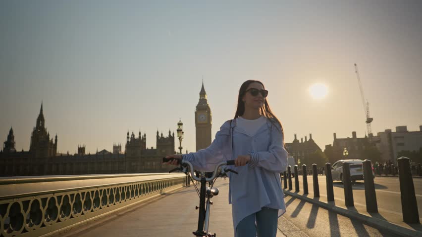 Stylish young tourist woman walking with bicycle on Westminster Bridge London with Big Ben view at sunset. Concept of eco-friendly transportation and healthy lifestyle in London Royalty-Free Stock Footage #1110165713