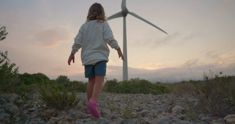 The cute little girl walking against at the sunset near the windmills. A child and a source of renewable ecological wind energy. Production of electricity in a wind farm