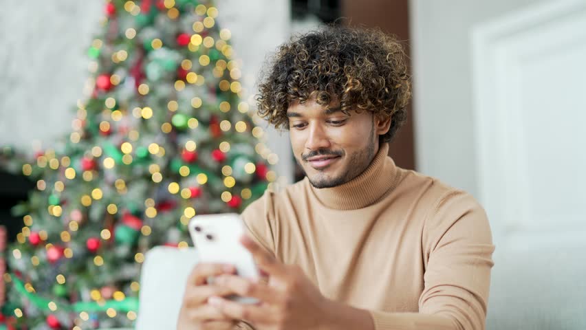 Close up. A young man uses a smartphone while sitting on sofa at home during winter New Year Christmas holidays. A handsome male is writing a greeting message, chatting with friends or shopping online Royalty-Free Stock Footage #1110169505
