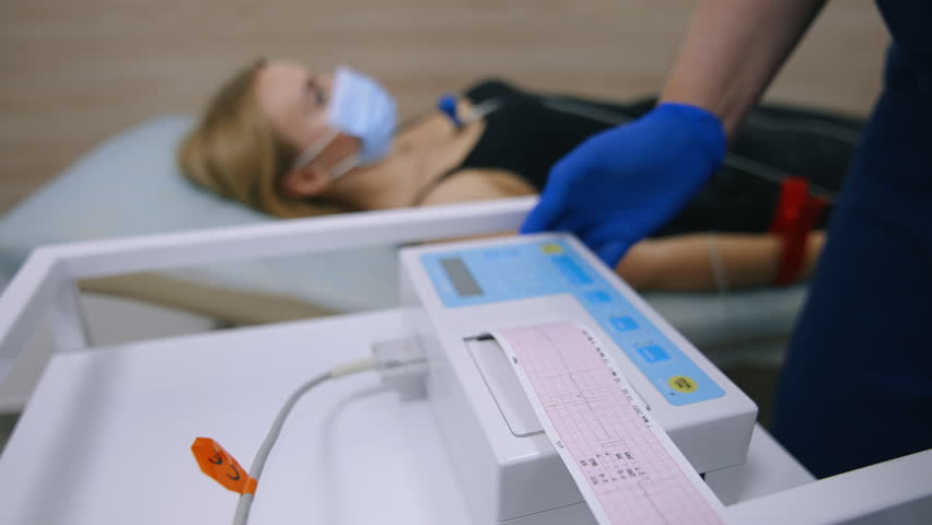 Paper with heart test result appears from electrocardiograph. Close up. Medic's gloved hand tears the paper from machine. Female patient at backdrop in blur. Royalty-Free Stock Footage #1110173229