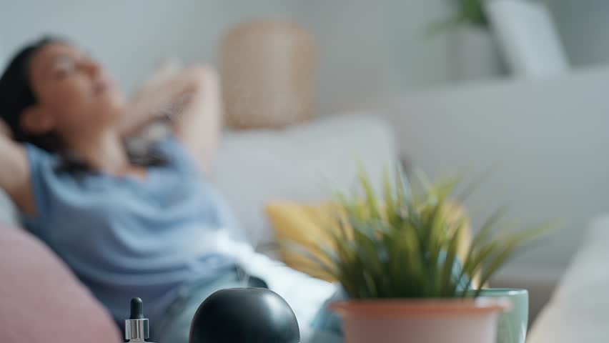 Video of beautiful woman relaxing on a coach while essential oil aroma diffuser humidifier the air in living room at home Royalty-Free Stock Footage #1110176269