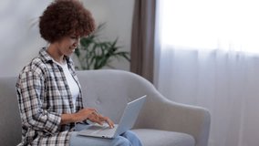 Happy hipster African American gen z woman, female student with afro hair talking with friend using laptop computer video conference calling in virtual webcam online chat sitting on sofa at home