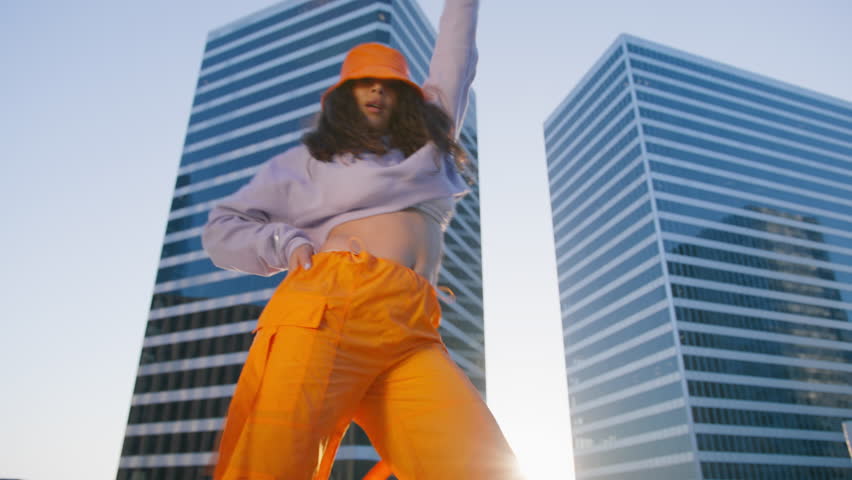 Low angle cinematic shot dancing woman young talented street style dancer performing freestyle hip hop moves in city. Mixes race model enjoying modern dance. Trendy lifestyle, cool expression close up