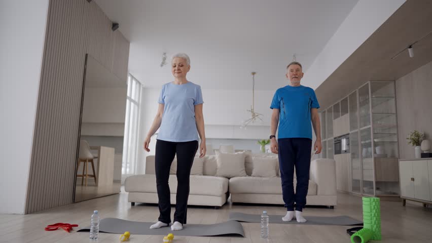 Positive senior couple man woman practicing yoga at home standing on mats. Happy wife husband doing static tree poses balancing on one leg falling. Sport, leisure, active healthy lifestyle concept. Royalty-Free Stock Footage #1110189045