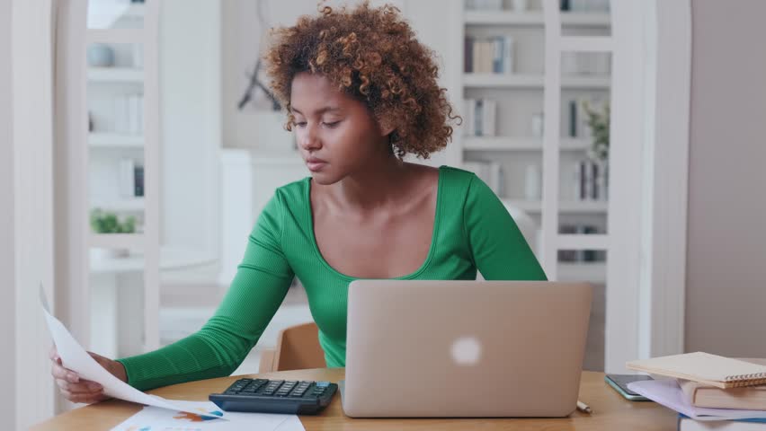 Young pretty African American woman financial analyst works from home studying documents with payment schedules and developing new economic strategy for company sits at table with laptop and books. Royalty-Free Stock Footage #1110189935