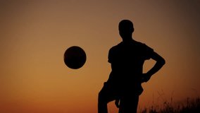 Medium full video capturing a silhouette of a teenager, young man playing with a ball on a field at sunset. He is kicking the ball in the air.