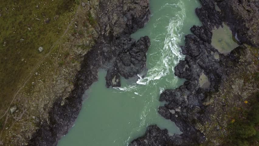Real time aerial view of brisk flowing deep river water through stony black mountainous terrain with hanging bridge parked vehicle near roadway green trees of Altai Republic Russia in daylight
