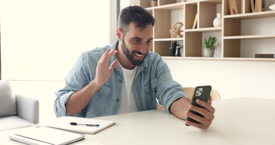 Young Hispanic guy holds cellphone makes video call sit workplace desk, smile looks at gadget screen greeting friend or family, share news, enjoy pleasant personal talk use videoconference application Royalty-Free Stock Footage #1110213561