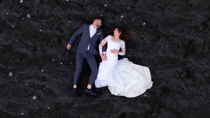 Drone footage of happy newlyweds having rest, hand in hand with one another, kissing on black sand of volcanic origin. Silhouettes recede and become tiny. High quality 4k footage Royalty-Free Stock Footage #1110218149