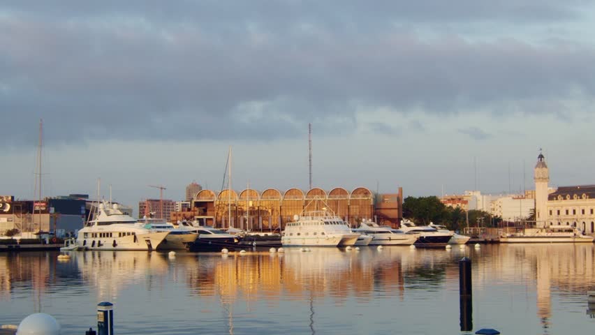 View of modern port with moored boats in sunlight.
Real time of yachts moored on calm river in harbor near town buildings under sky with clouds at sundown