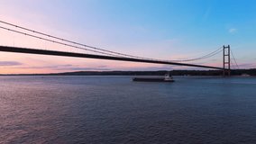 An unhurried barge glides beneath Humber Bridge at sunset, captured in a mesmerizing aerial drone clip during the golden hour.