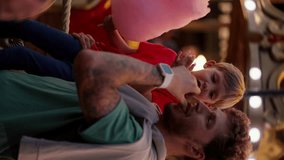 Vertical video of a father and his little blond son with blue hair in a red T-shirt eating pink cotton candy in Luna Park in the evening