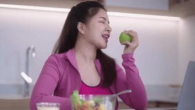 sporty young Asian woman in sportswear having an online video call via laptop while eating healthy organic apple fruit doing fitness training recommending healthy nutrition in the kitchen at home