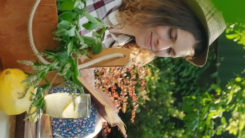 Vertical shot. Young woman reading book drinking refreshing lemonade cocktail siting at table in summer garden. Royalty-Free Stock Footage #1110236367