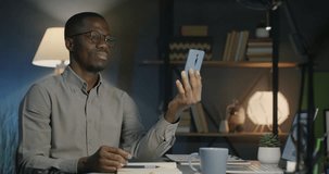 African American man entrepreneur making online video call using smartphone messenger in dark office in evening. People and professional communication concept.