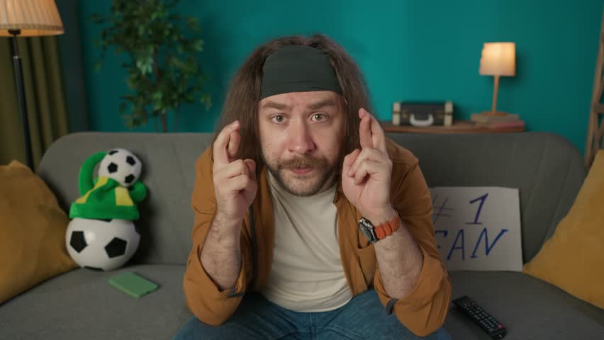 The frame shows a middle aged man sitting on a sofa. Demonstrates watching a soccer match. The camera is distant, he has his fingers crossed and hopes for a win. He then rejoices, kissing the ball