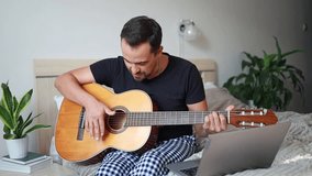 A man learns to play the guitar by watching instructional videos on a laptop at home on his bed.