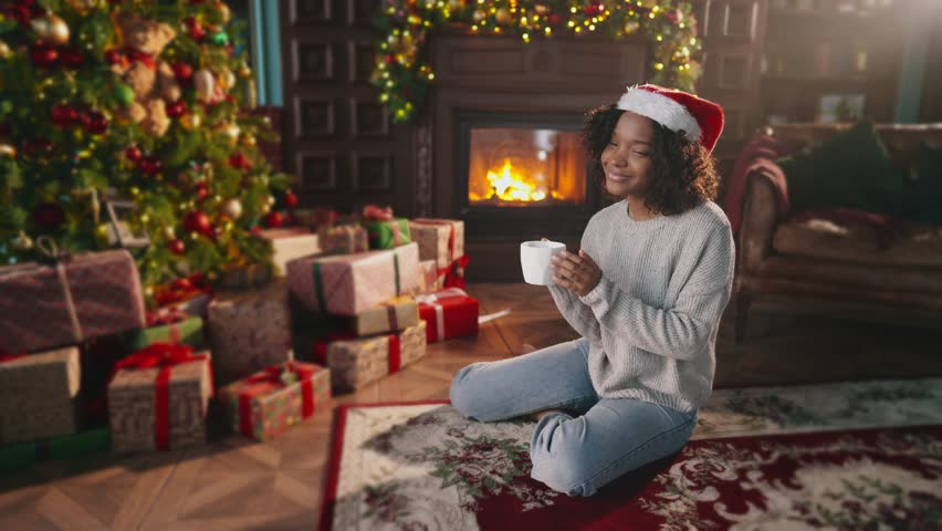 Xmas scene. Christmas, smile or happy african american woman with tea in home on holiday. Beauty young Christmas woman sitting near fireplace in dark room at home and drinking hot beverage.