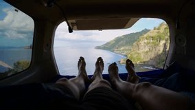Young traveler couple laying in camping trailer bed, enjoying view from open window and swinging their feet. Cinematic view from cozy camping trailer to mountains and ocean. Travel by camping trailer.